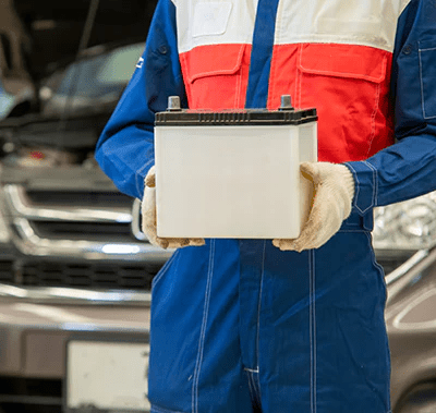 Person in a blue, orange, and white jumpsuit holding a white car battery standing in front of a car with a popped hood.