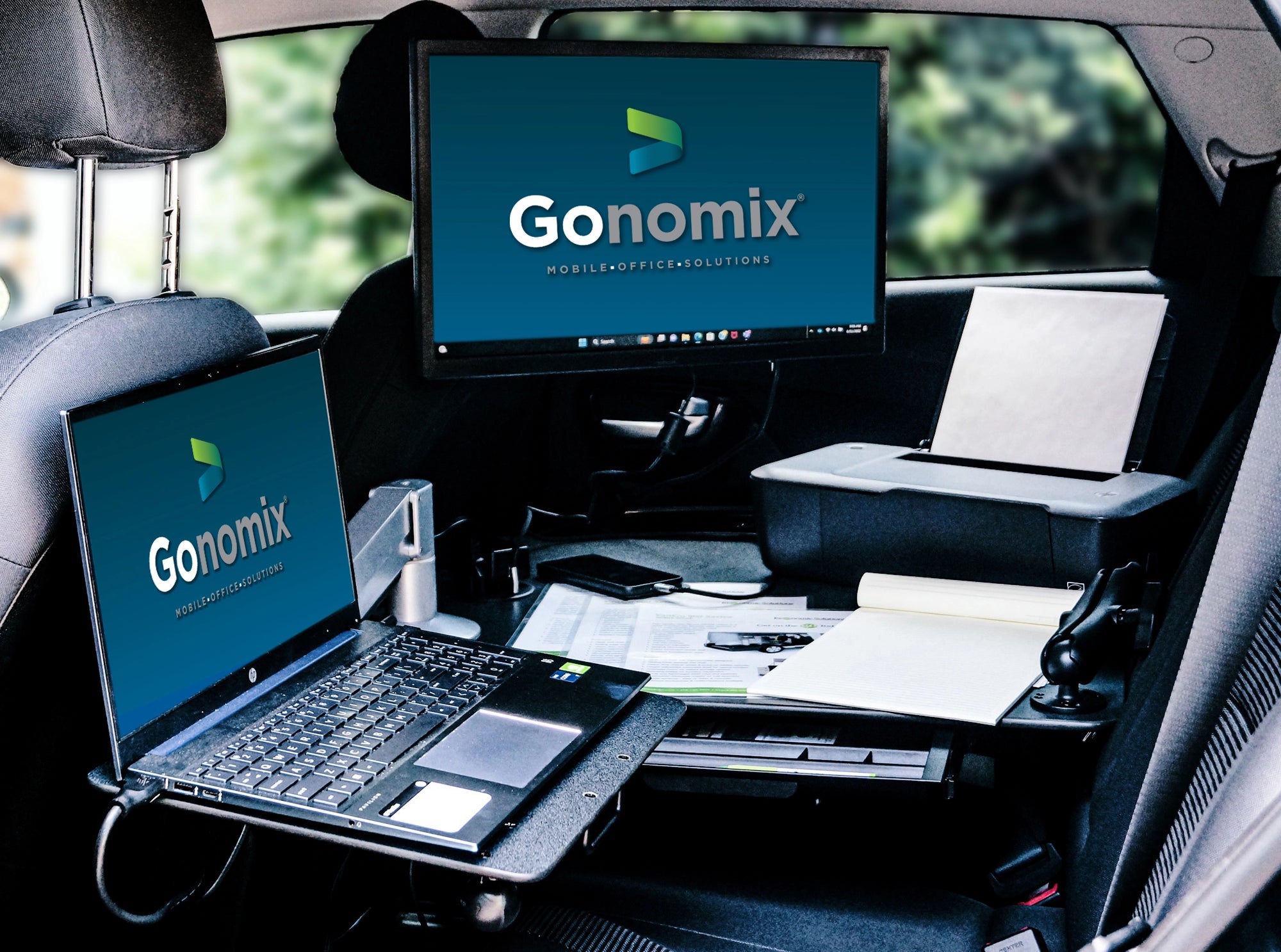 A mobile office set up inside a SUV with a sleek black desk buckled in the rear passenger seat. There is an articulating laptop mount, with a laptop,  positioned over the rear driver's seat and another monitor mounted by the back of the passenger's seat. The desk has a portable printer, papers, and a cellphone on the surface. The monitor and laptop screen depict a website with a large white van in the center. The vehicle is parked in an outdoor setting with greenery visible through the windows. 