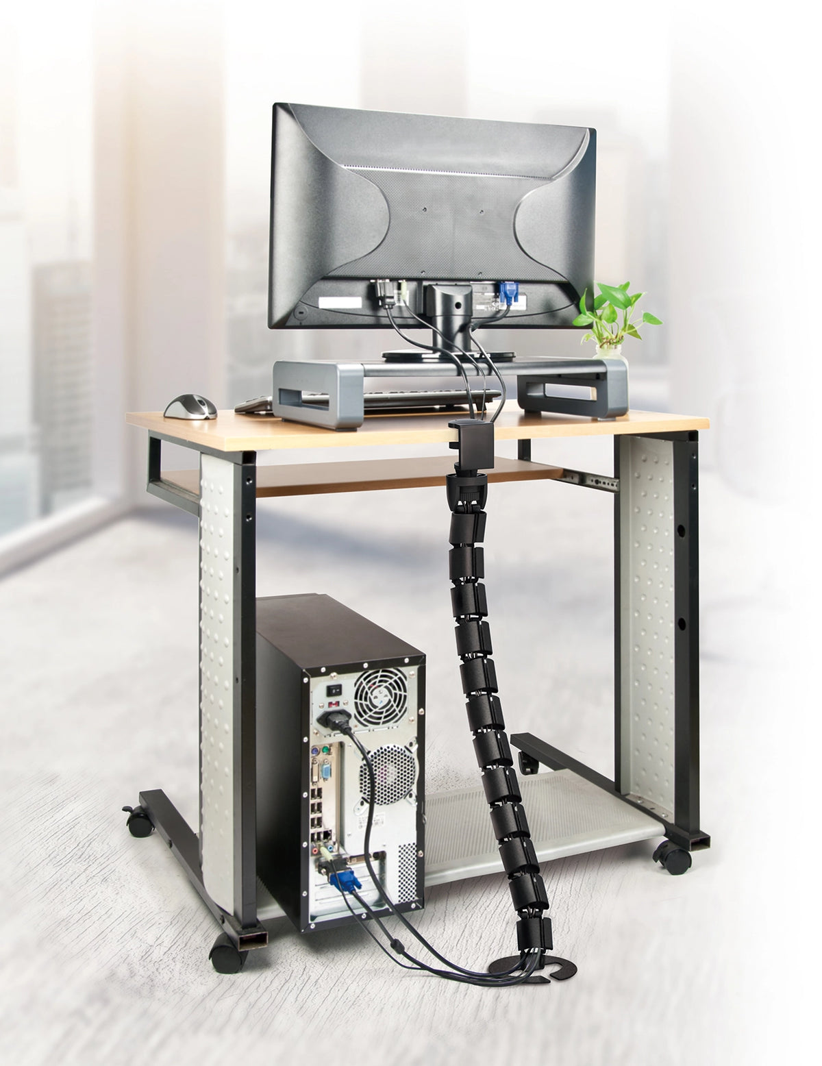 a vertical, segmented cable management spine attached to the underside of a desk. It’s black and has multiple segments, giving it a flexible, snake-like appearance. The spine is holding several cables, keeping them organized and preventing them from tangling. The desk setup includes a monitor, a small plant, an external hard drive, and a CPU tower