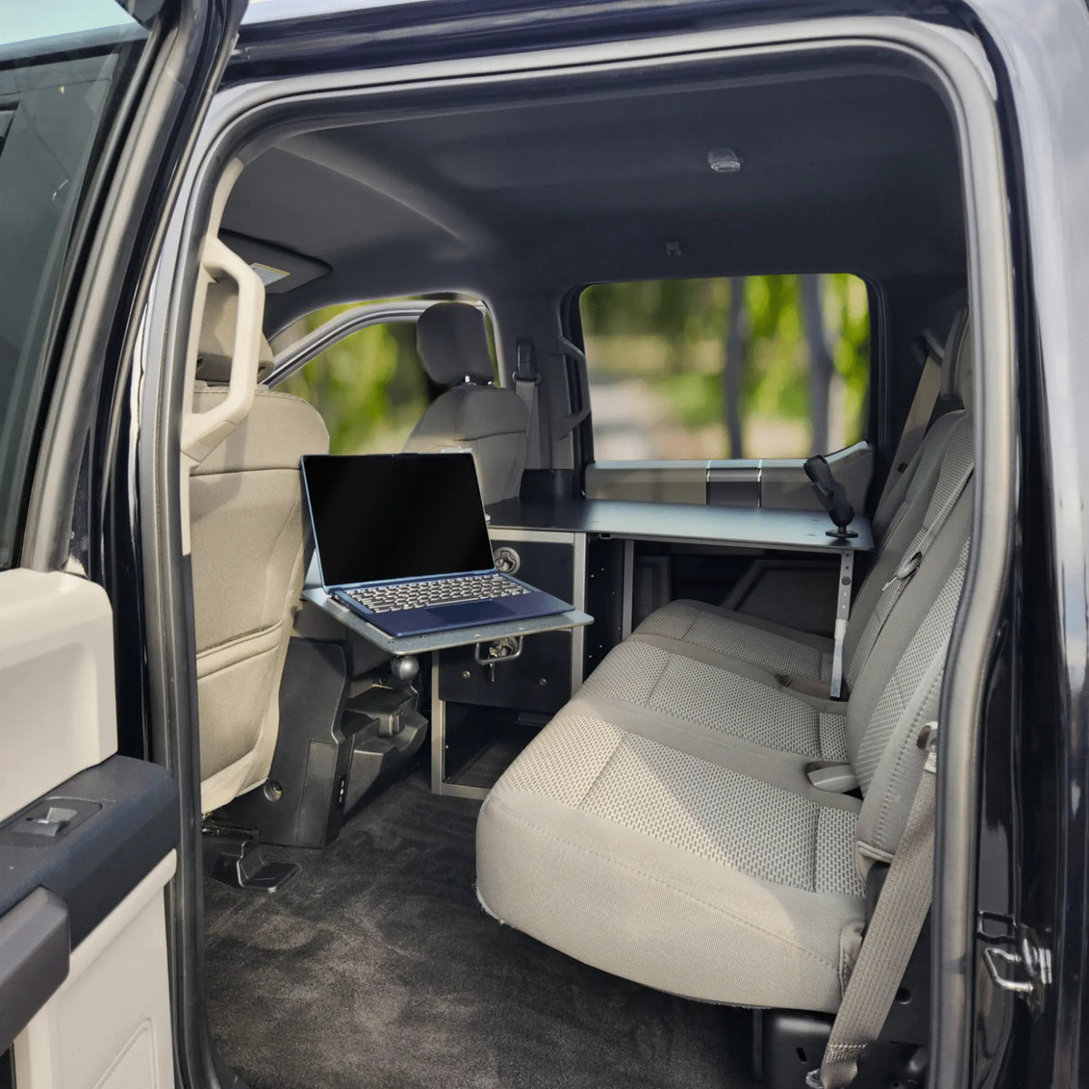 A mobile office set up inside a pickup truck with tan seats and a light interior. A sleek black desk is mounted and rests on the rear passenger seat. There is an articulating laptop mount, with a laptop, positioned over the rear driver&#39;s seat. The desk has a set of drawers located in the leg space of the rear passenger seat. The vehicle is parked in an outdoor setting with greenery visible through the windows. 