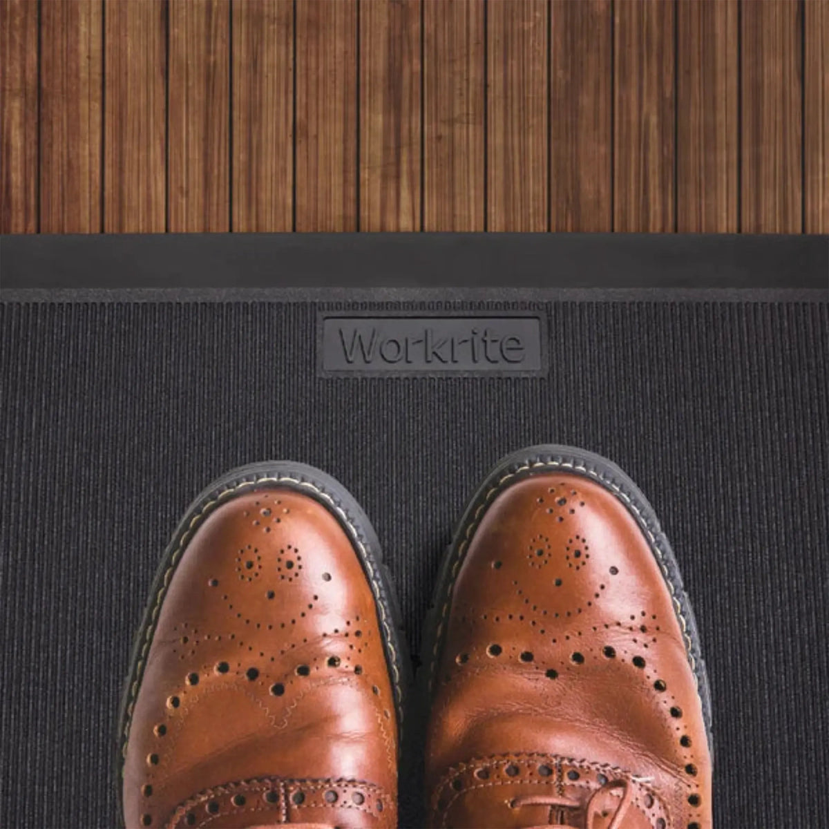 A pair of brown leather brogue shoes standing on a black Workrite anti-fatigue mat, with a wood plank floor visible above the mat