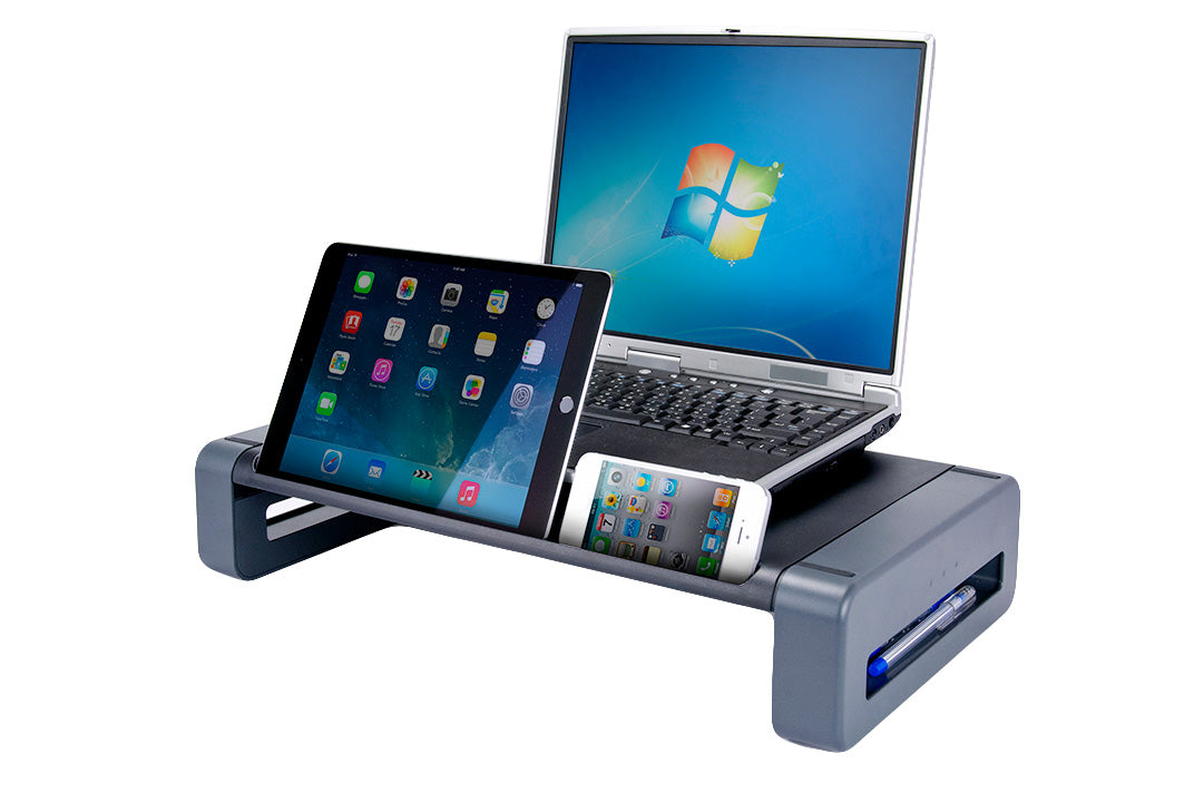 a rectangular monitor stand with a black surface tray and a gray stand. On the surface there is a long divot in which a vertical black iPad and white iphone rest. An open silver laptop rests on the main surface