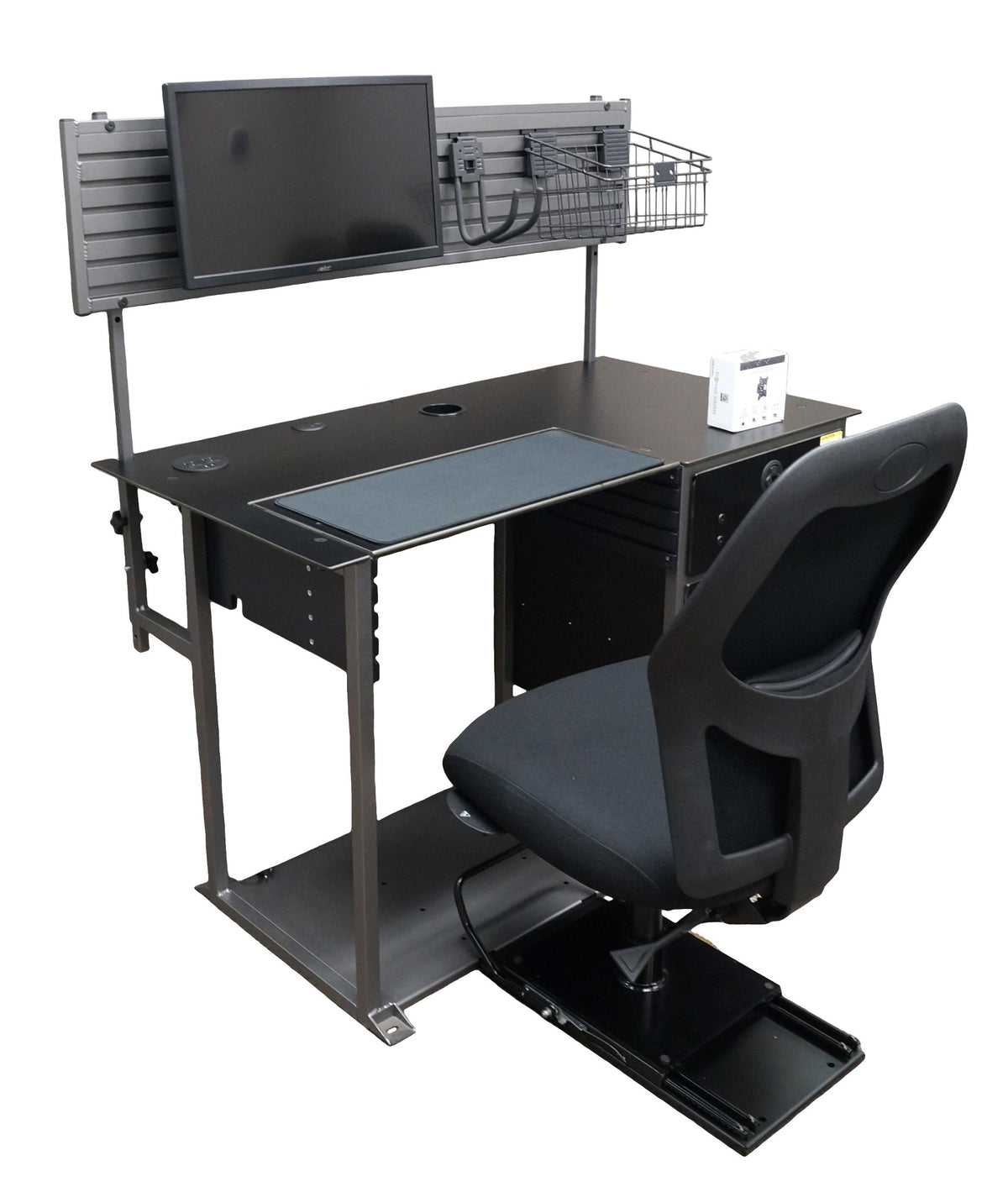 a right-angled view of a small black desk, a gray metal hutch with a monitor, hook, and basket, and a black chair mounted on a sliding seat base. It has a silver metal frame and a set of drawers on the right side. The work surface has 2 grommet holes, a white box, and a sheet of black fabric in the middle for a keyboard and mouse. The hutch is mounted to the desk with metal poles on the back. The bottom is connected together with a large sheet of gray metal to which the sliding black chair is mounted. 