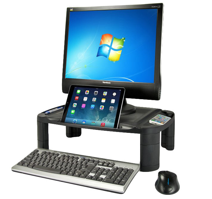 a rectangular, black plastic monitor stand with a textured top surface to support a monitor. There is a long divot intended for phones, tablets, pens, etc. The stand has four cylindrical legs at each corner. There is a large flatscreen monitor, a black iPad in the long divot at the front, and a white iphone resting on the right-side surface. At the base of the stand there is a standard silver keyboard and black ergonomic mouse