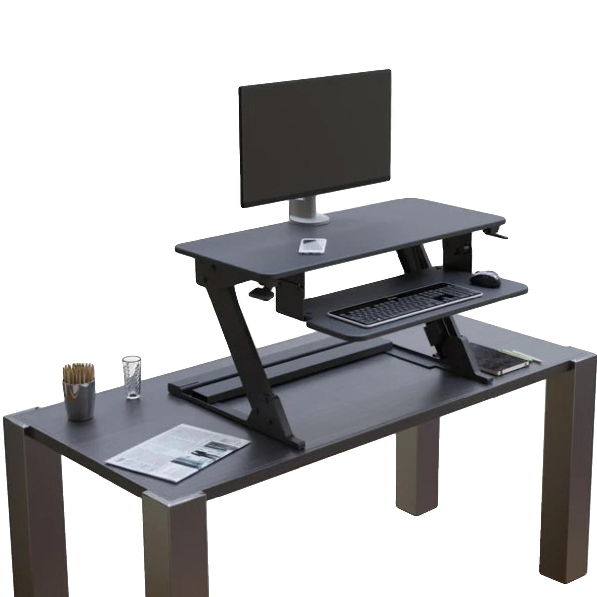 a desktop standing desk converter on a standard desk, featuring a two-tiered design with an upper display surface for a monitor and a lower retractable keyboard and mouse tray. The setup includes a single monitor, pencil holder, glass of water, and documents on the main desk